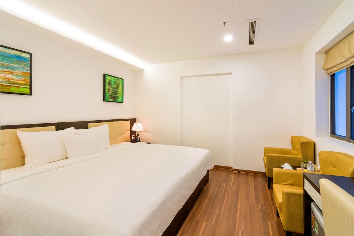 Quarto de hotel com uma cama grande, uma mesinha de cabeceira com abajur, uma janela com vista e um espaço de estar com cadeiras.