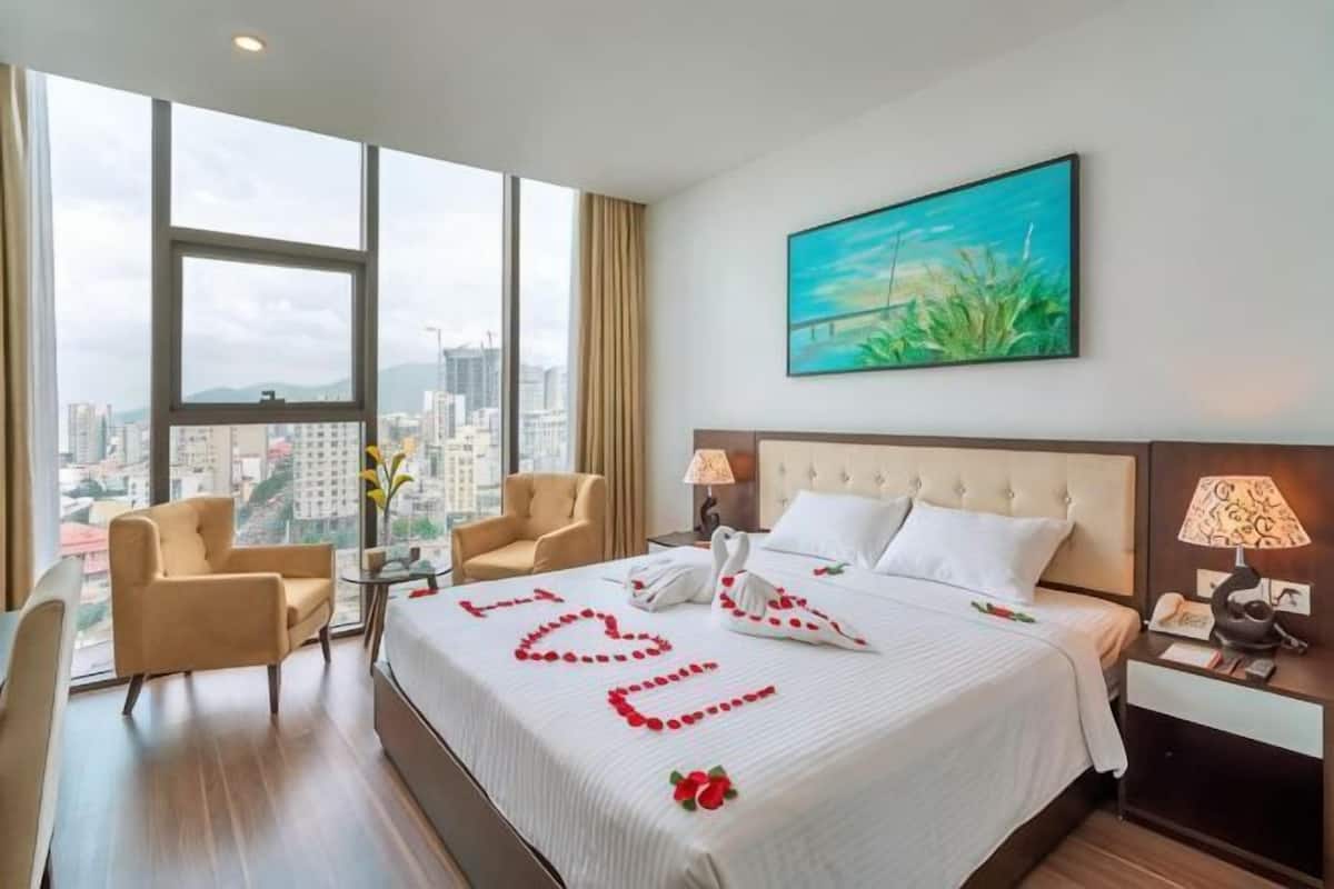 Habitación de hotel con una cama grande, dos mesitas de noche, vistas a la ciudad y un cuadro en la pared.