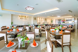 A restaurant with multiple dining tables set with orange napkins and green plants. - Dana Marina Hotel (Da Nang)