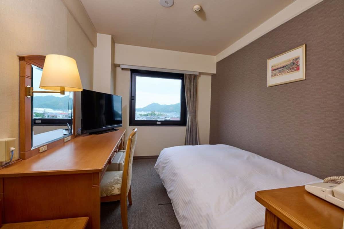 Une chambre d’hôtel avec un lit, un bureau avec une lampe, une chaise, une télévision, une fenêtre donnant sur les montagnes et un tableau encadré au mur.