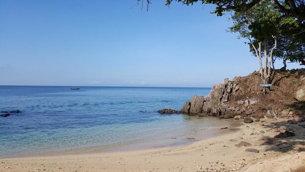 Una playa con agua azul cristalina, un barco a lo lejos y terreno rocoso con árboles.