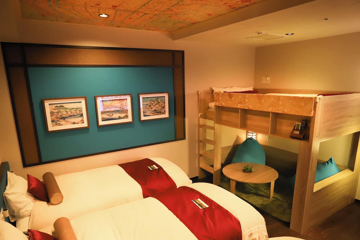 A hotel room with two beds, a bunk bed, a small table, and a wall with framed pictures.