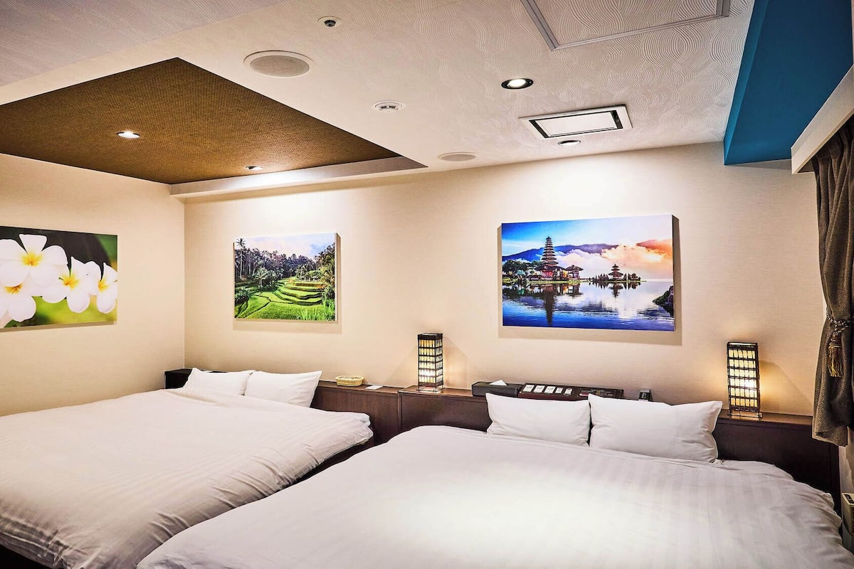 A hotel room with two beds, a ceiling with recessed lighting, and three wall-mounted paintings depicting flowers, a landscape, and a temple.