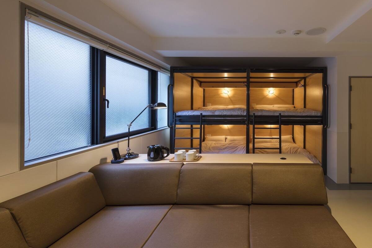 A compact room with a bunk bed, a sofa, a desk with a lamp, and a window with blinds.