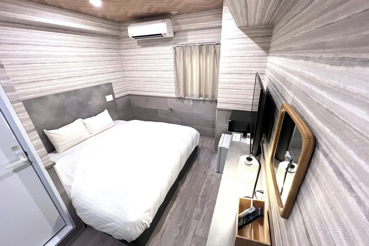 A modern hotel room with a large bed, a bedside table with a lamp, a mirror, and an air conditioning unit.