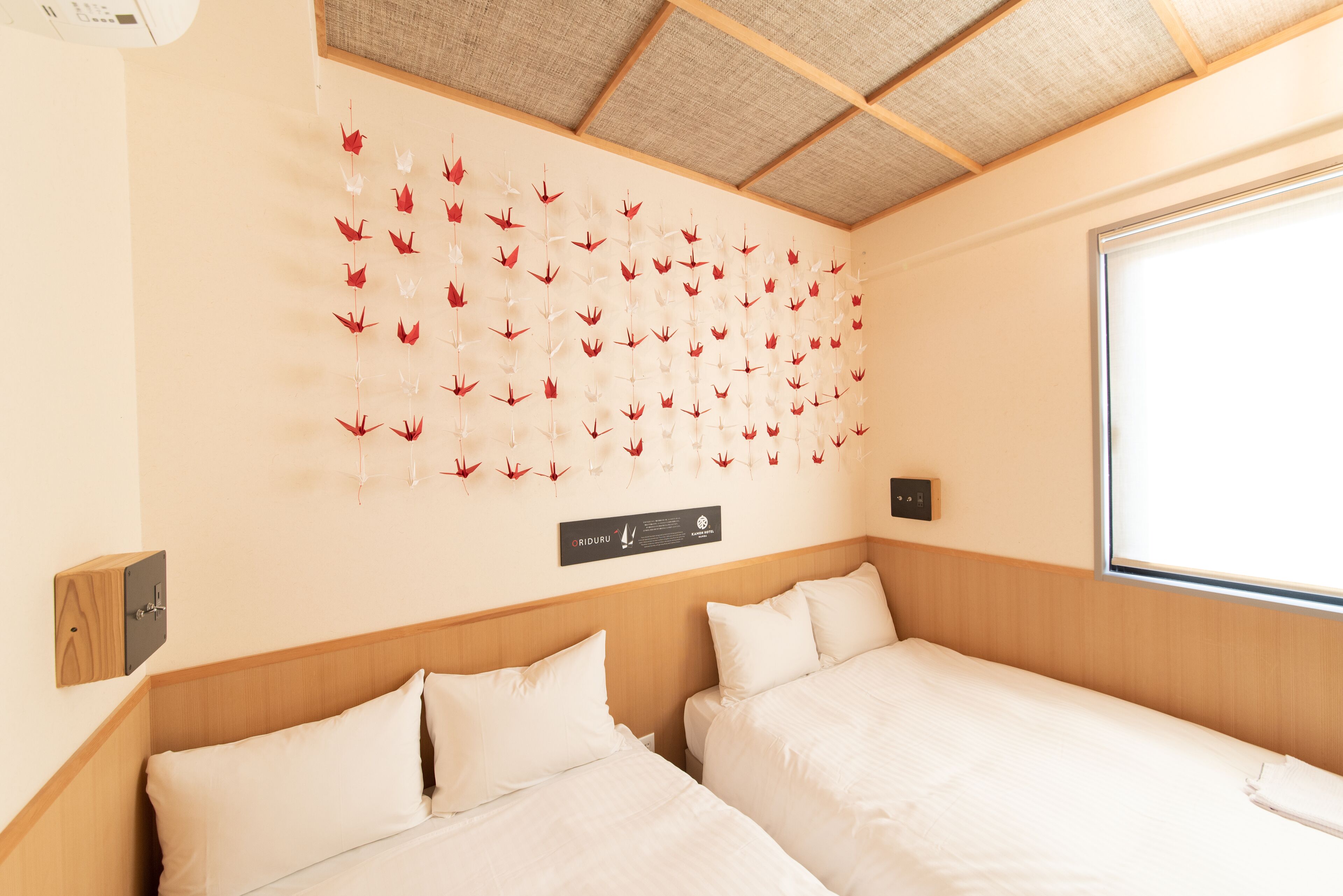 A hotel room with two beds, a wooden headboard, a wall-mounted artwork of red and white leaves, and a window with blinds.