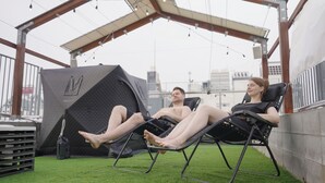 Twee personen ontspannen in loungestoelen in een buitengebied met kunstgras, een tent en een stadsgezicht op de achtergrond.