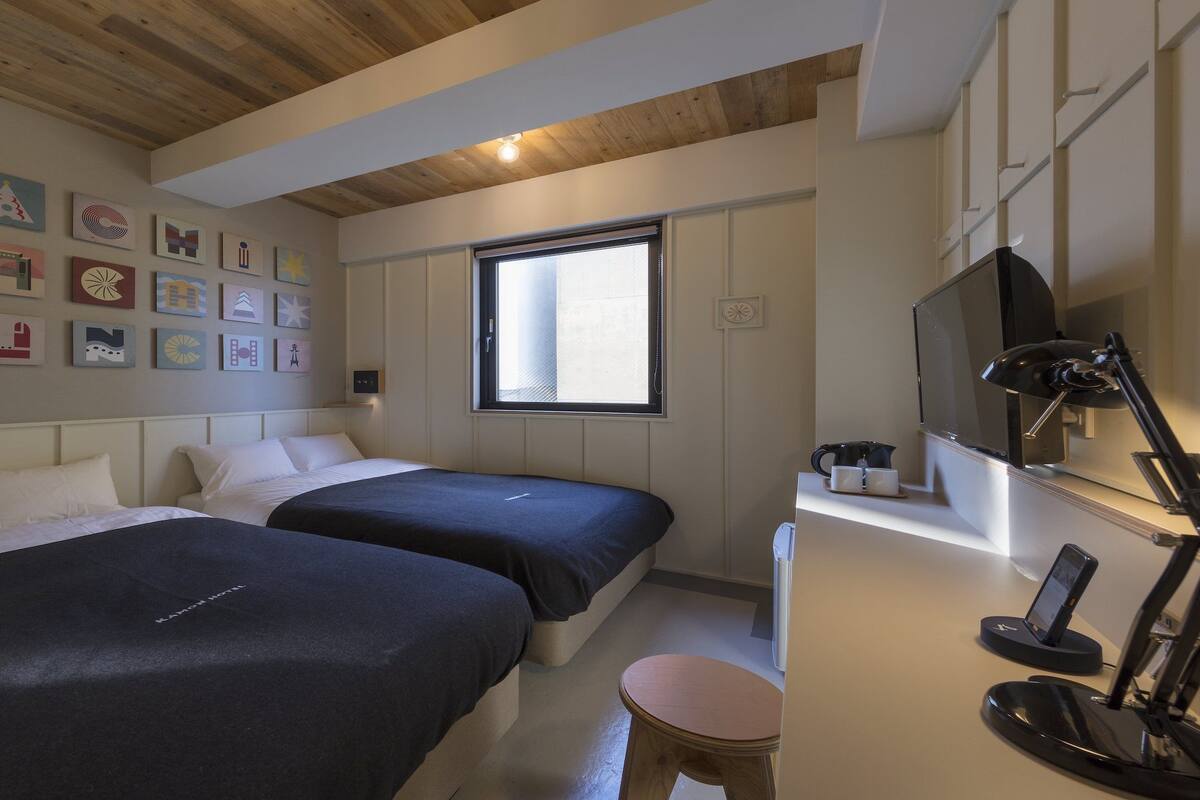 A bedroom with two beds, a desk with a computer, a TV, and a window with blinds.