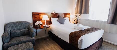 Una habitación de hotel con una cama, una silla, una mesita de noche con lámpara y una ventana con cortinas.