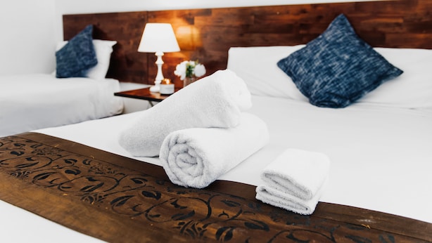 A neatly made bed with white linens and rolled towels, a wooden headboard, and a nightstand with a lamp and a small vase with flowers. - Tamworth City Motel (South Tamworth)