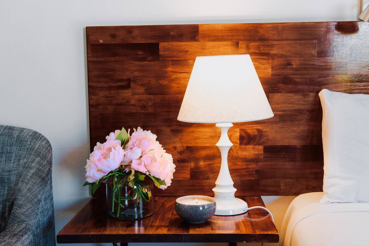 Une tête de lit en bois, une lampe blanche, un vase avec des fleurs roses et un petit bol posé sur une table de chevet en bois.