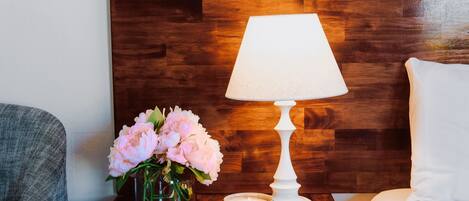 Un cabecero de madera, una lámpara blanca, un jarrón con flores rosadas y un pequeño cuenco sobre una mesita de noche de madera.
