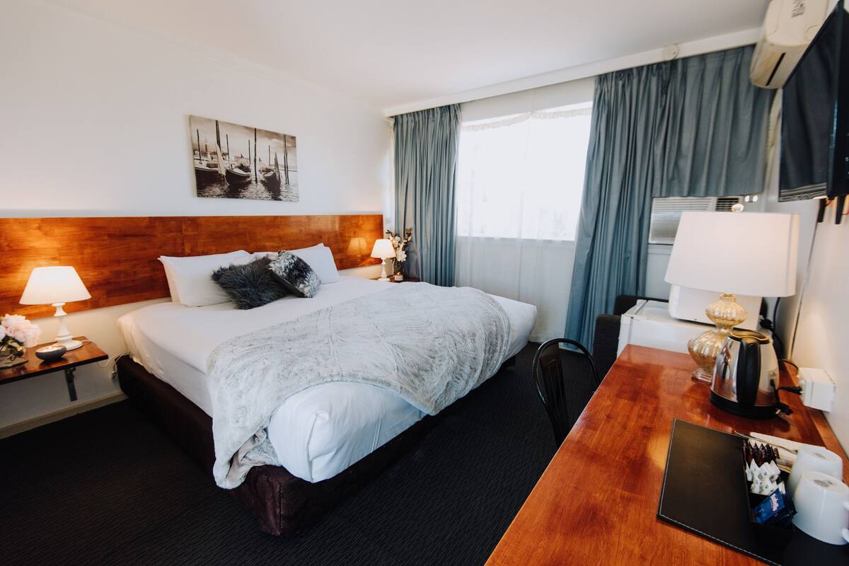 Une chambre d’hôtel avec un grand lit, deux tables de chevet avec des lampes, un bureau avec un ordinateur portable, une machine à café et un climatiseur fixé au mur.