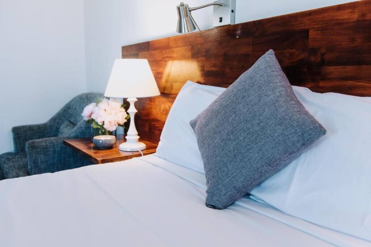 Un lit bien fait, avec un oreiller gris, une lampe blanche posée sur une table de chevet en bois, et un vase rempli de fleurs.