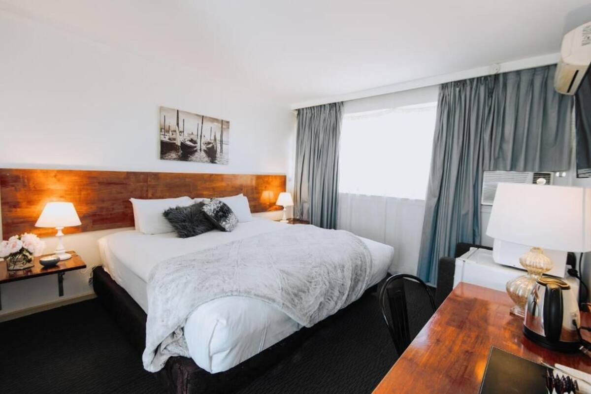Une chambre d’hôtel avec un grand lit, deux lampes de chevet, une tête de lit en bois, un tableau, un bureau avec une lampe et une fenêtre avec des rideaux.