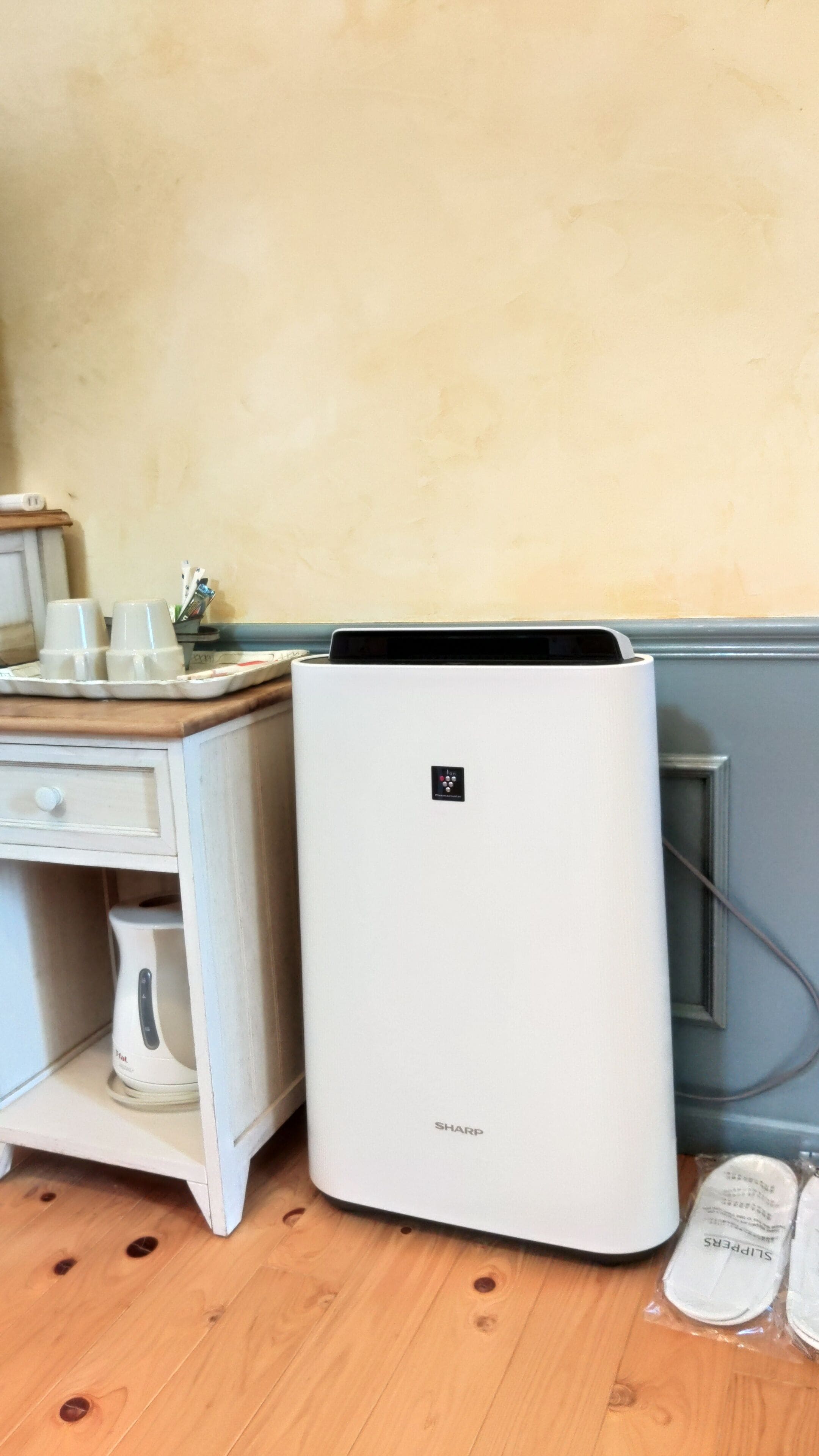 A white air conditioner unit with a control panel, placed on a wooden floor next to a dresser with a mirror and a cup holder.
