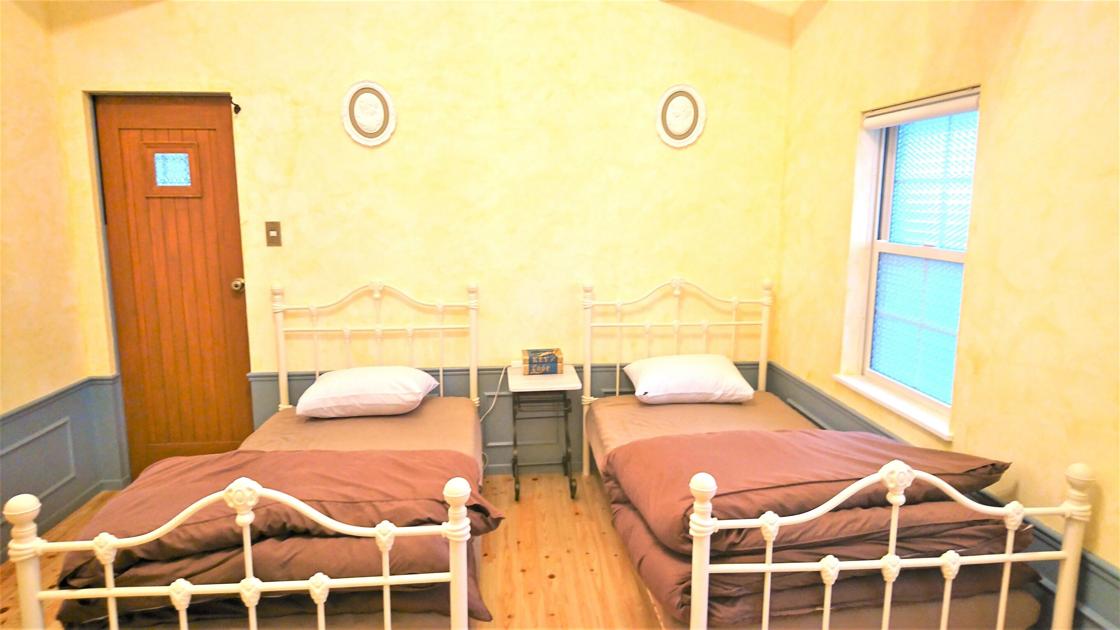 A room with two beds, a wooden door, a small table with a book, and a window.
