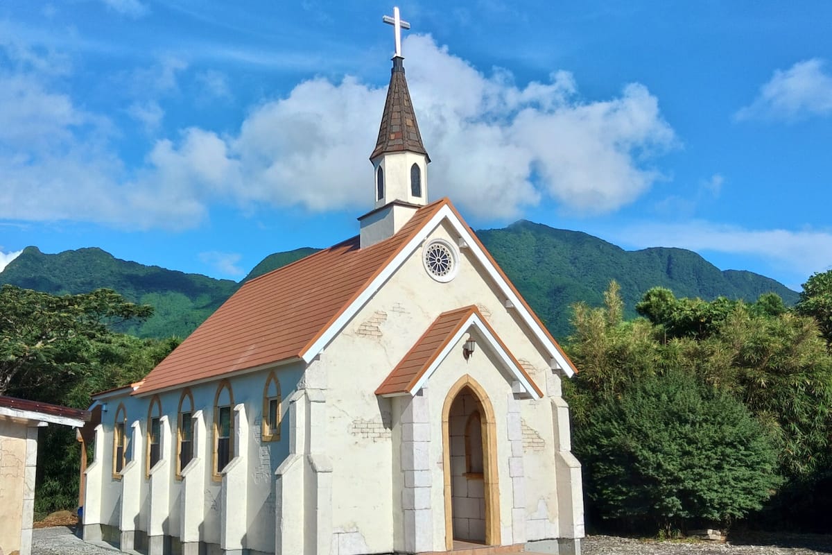 A church with a steeple and a cross, surrounded by greenery and mountains.