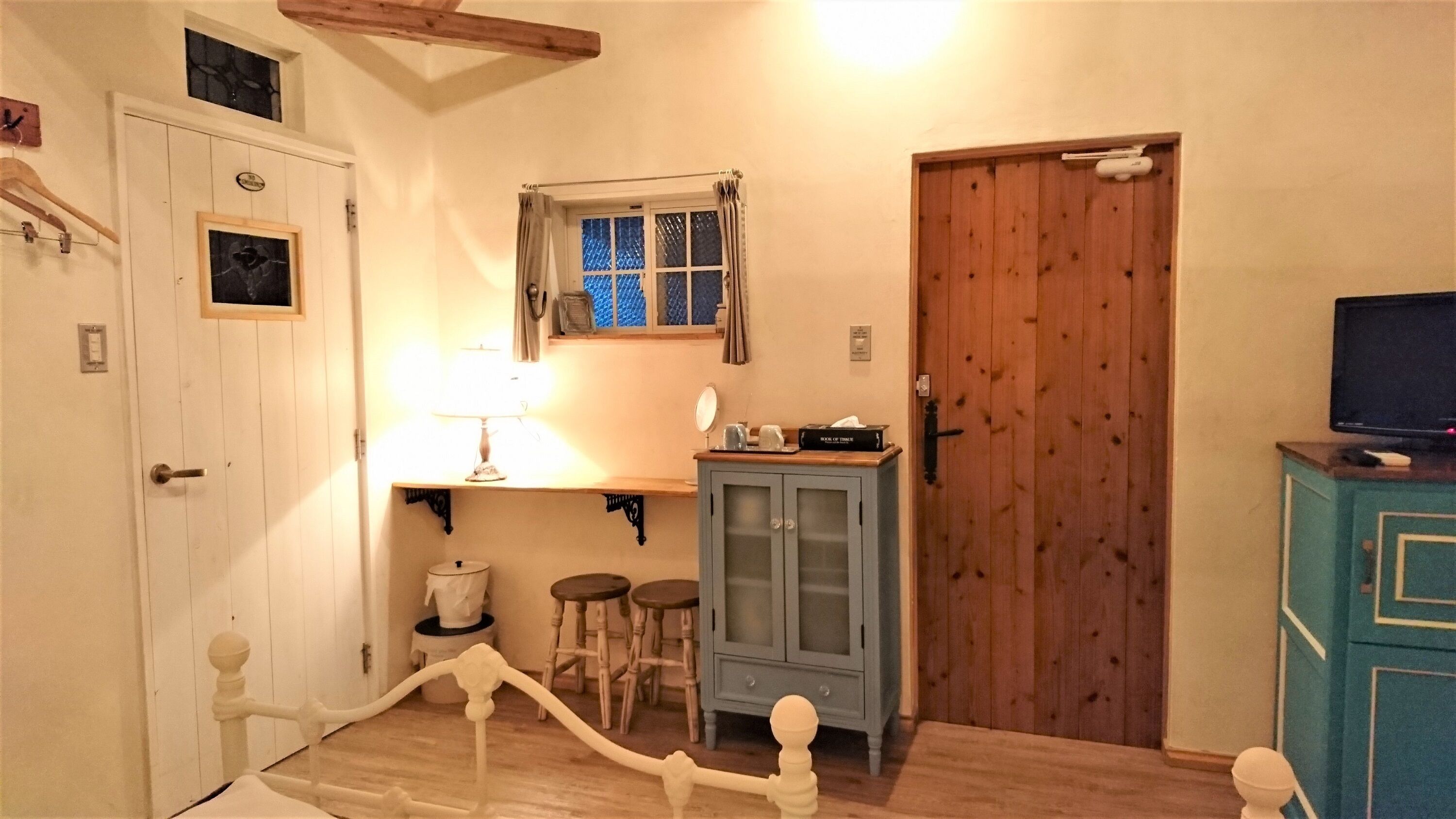 Una habitación con puerta de madera, un ventanal pequeño con cortinas, un armario de madera, una mesa con lámpara y un televisor sobre un mueble.