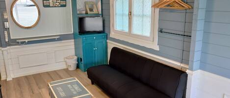 A room with a black sofa, a small table, a teal cabinet, a mirror, and a window with a wooden hanger rack.