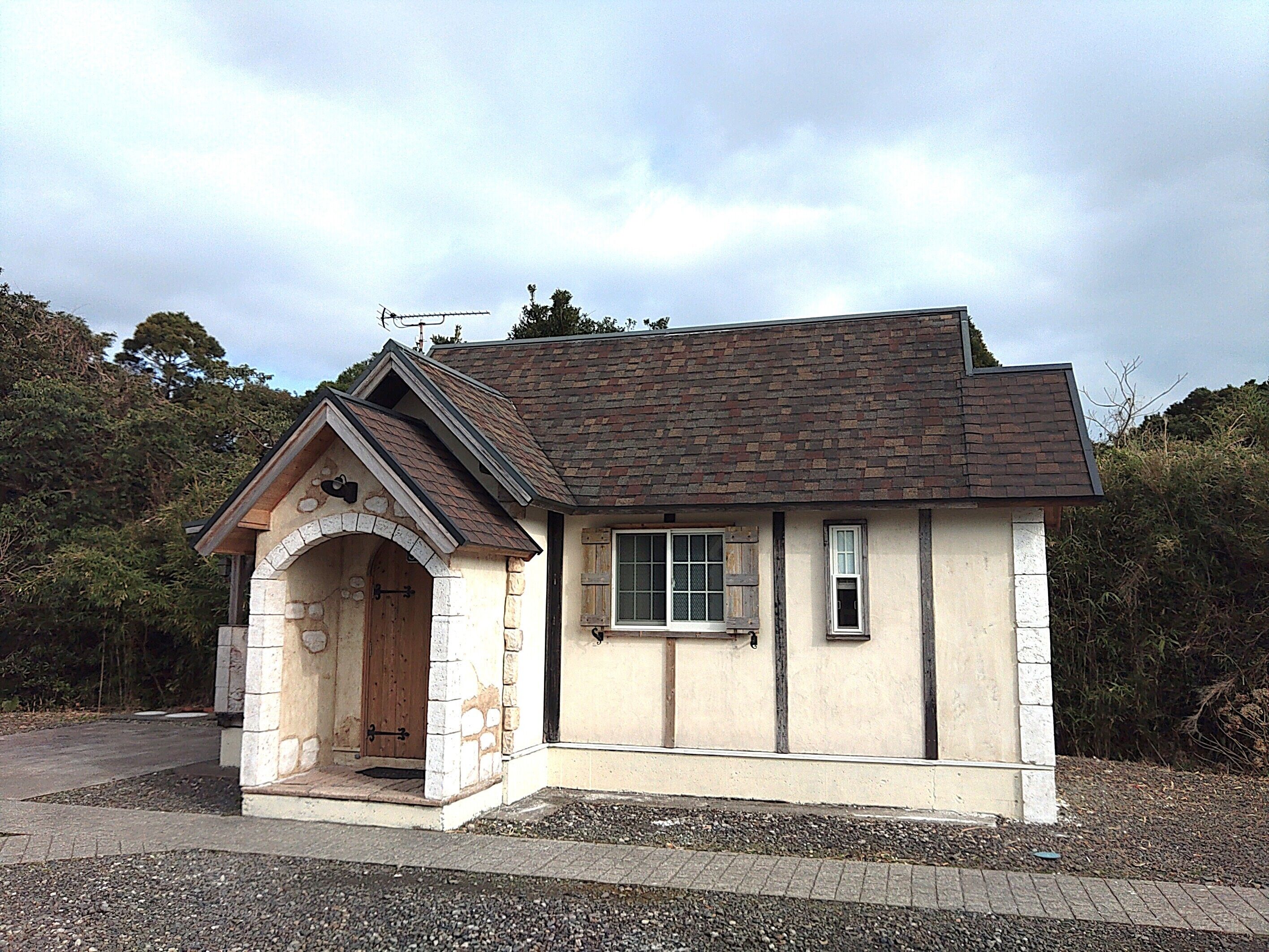 A small, single-story house with a stone archway entrance, a pitched roof, and a window with shutters.