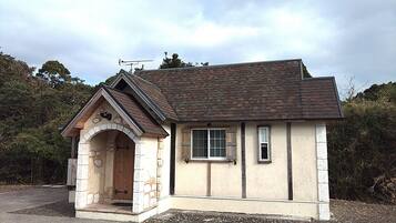 A small, single-story house with a stone archway entrance, a pitched roof, and a window with shutters.