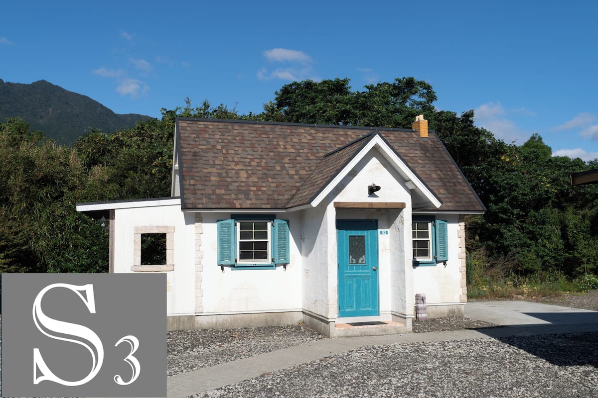 A small white building with a blue door and shutters, surrounded by gravel and greenery.
