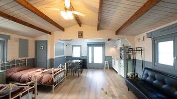 A room with a bed, a sofa, a dining table with chairs, a kitchen area, and a ceiling fan.