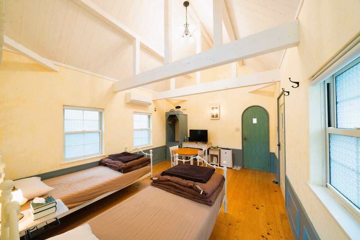 A dormitory room with two single beds, a desk with a chair, a television, and a window.