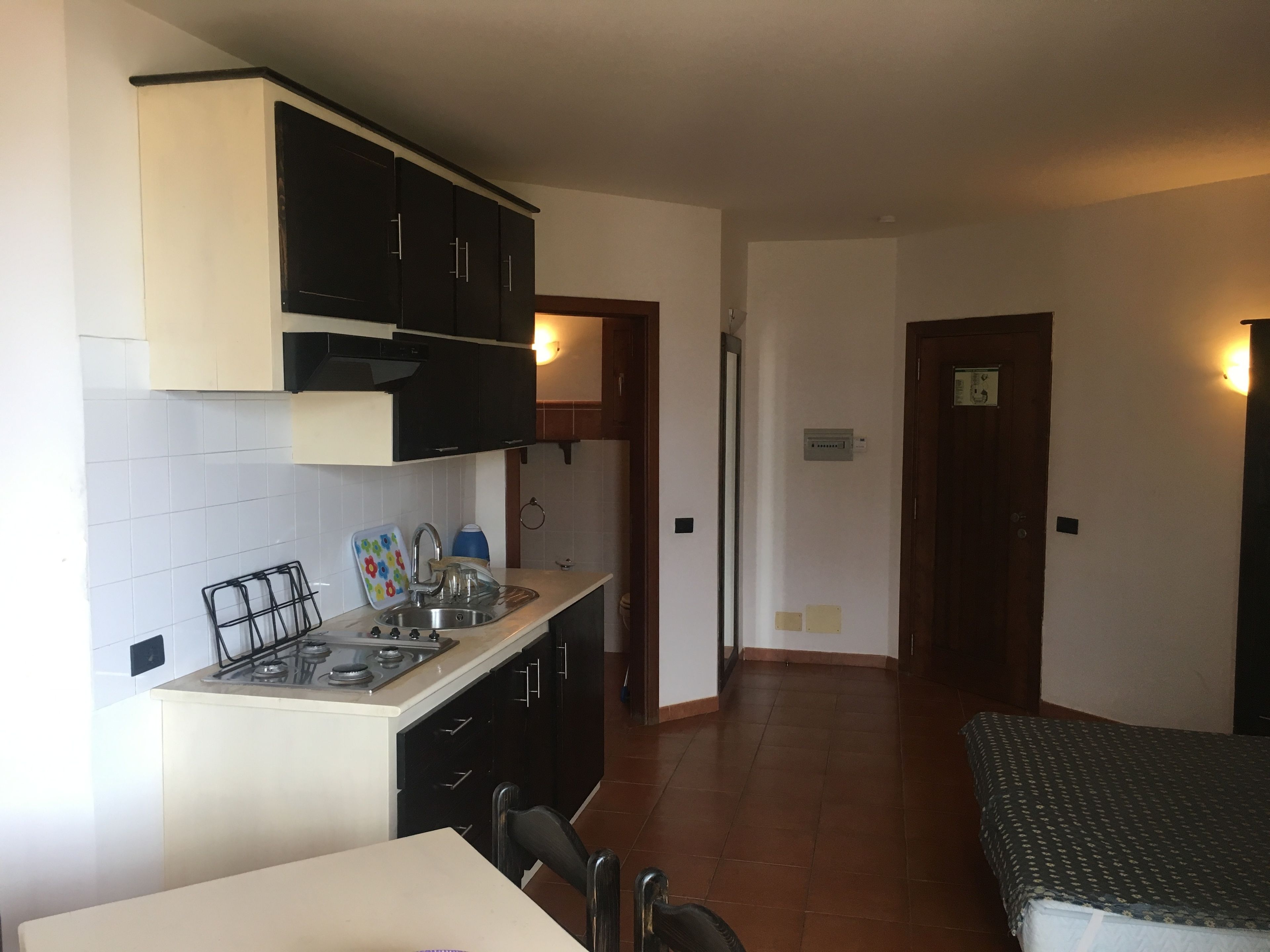 A compact kitchen with a stove, sink, and cabinets. There is a dining table with chairs and a bed in the adjacent room.
