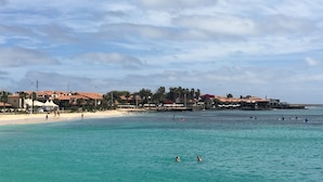 A coastal beach scene with clear turquoise waters, a sandy shore, and buildings with red roofs in the background. - Porto Antigo (Sal)