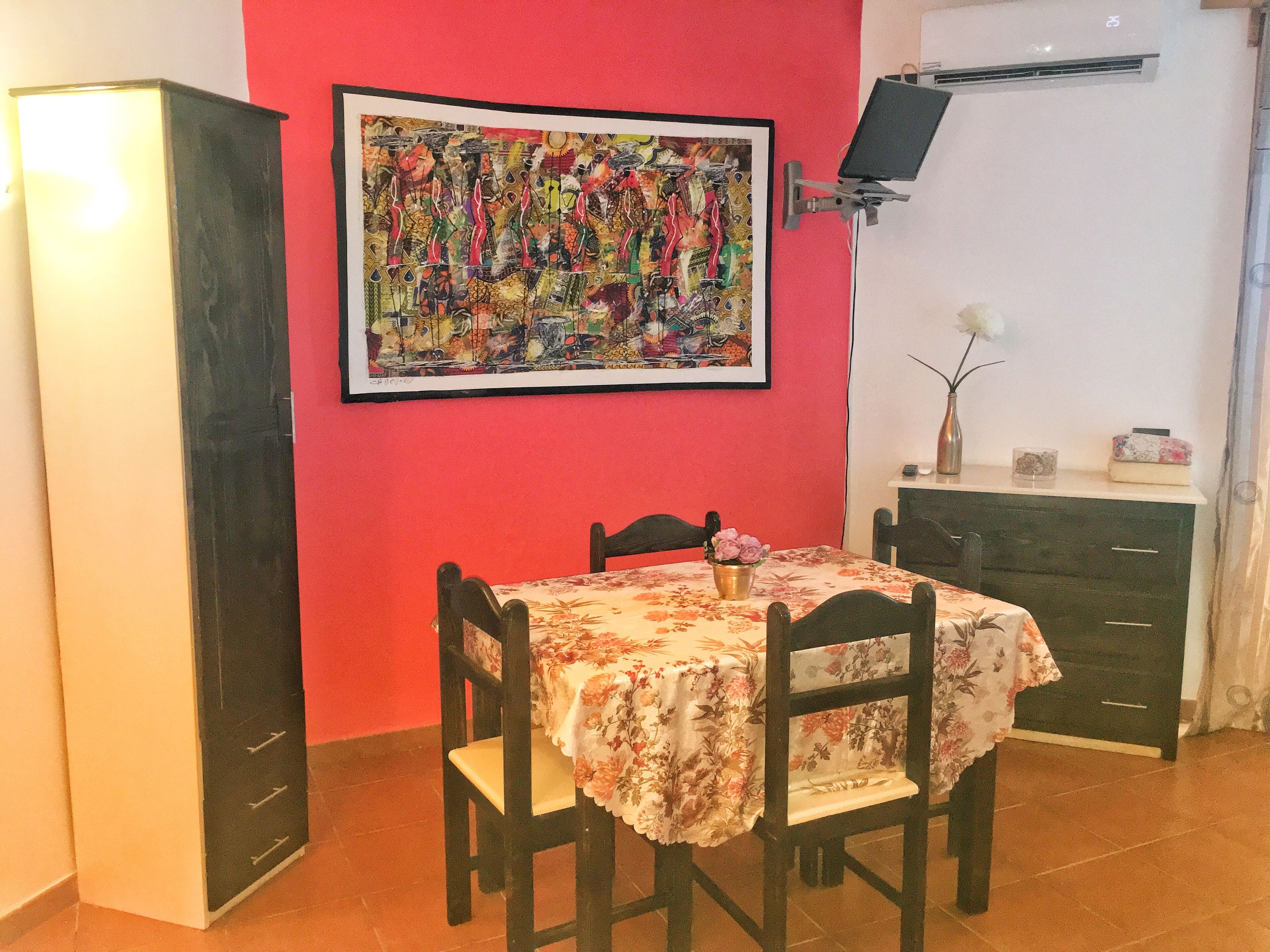 Un comedor con una mesa preparada para dos, una pared roja, un cuadro abstracto enmarcado y un televisor montado en la pared.