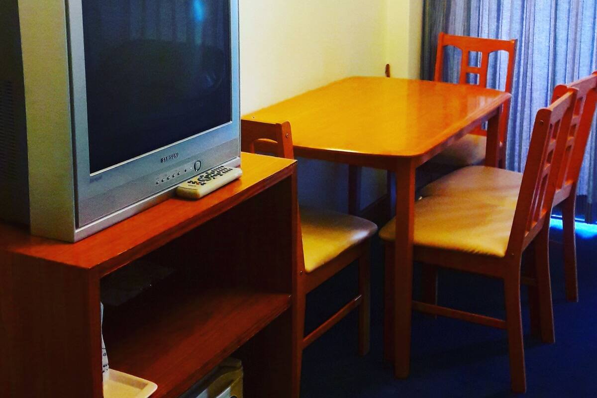 Un téléviseur posé sur un meuble en bois, une table en bois et deux chaises en bois dans une pièce avec un rideau et un interrupteur sur le mur.