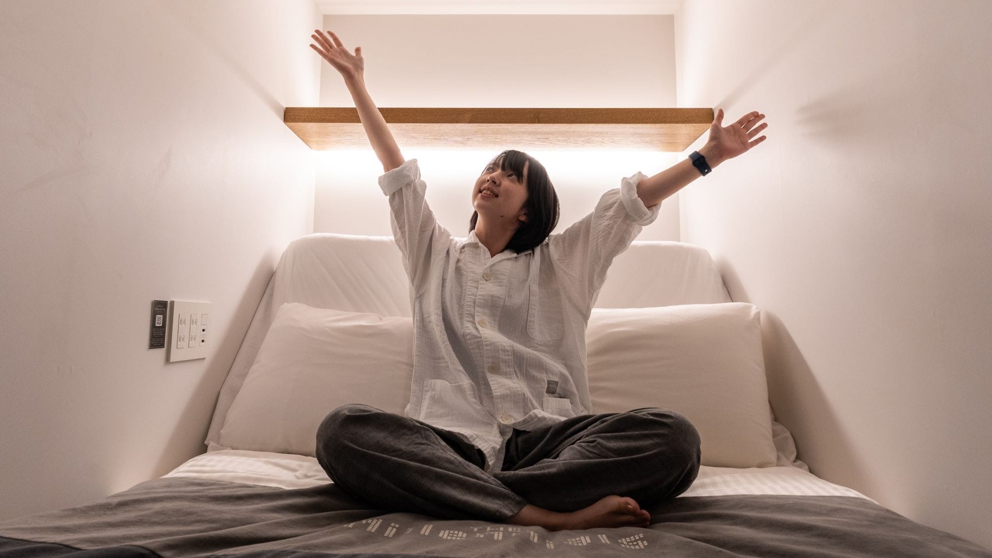 Una persona sentada en una cama con un almohadón blanco y un cabecero de madera, en una habitación con pared blanca y un interruptor de luz a la vista.
