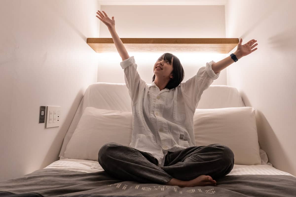 Uma pessoa sentada em uma cama com travesseiro branco e cabeceira de madeira, em um quarto com parede branca e um interruptor de luz visível.