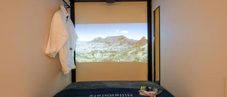 Un dormitorio con cama, una pantalla de proyector mostrando un paisaje montañoso, una toalla colgada en un perchero y una mesita con libros.