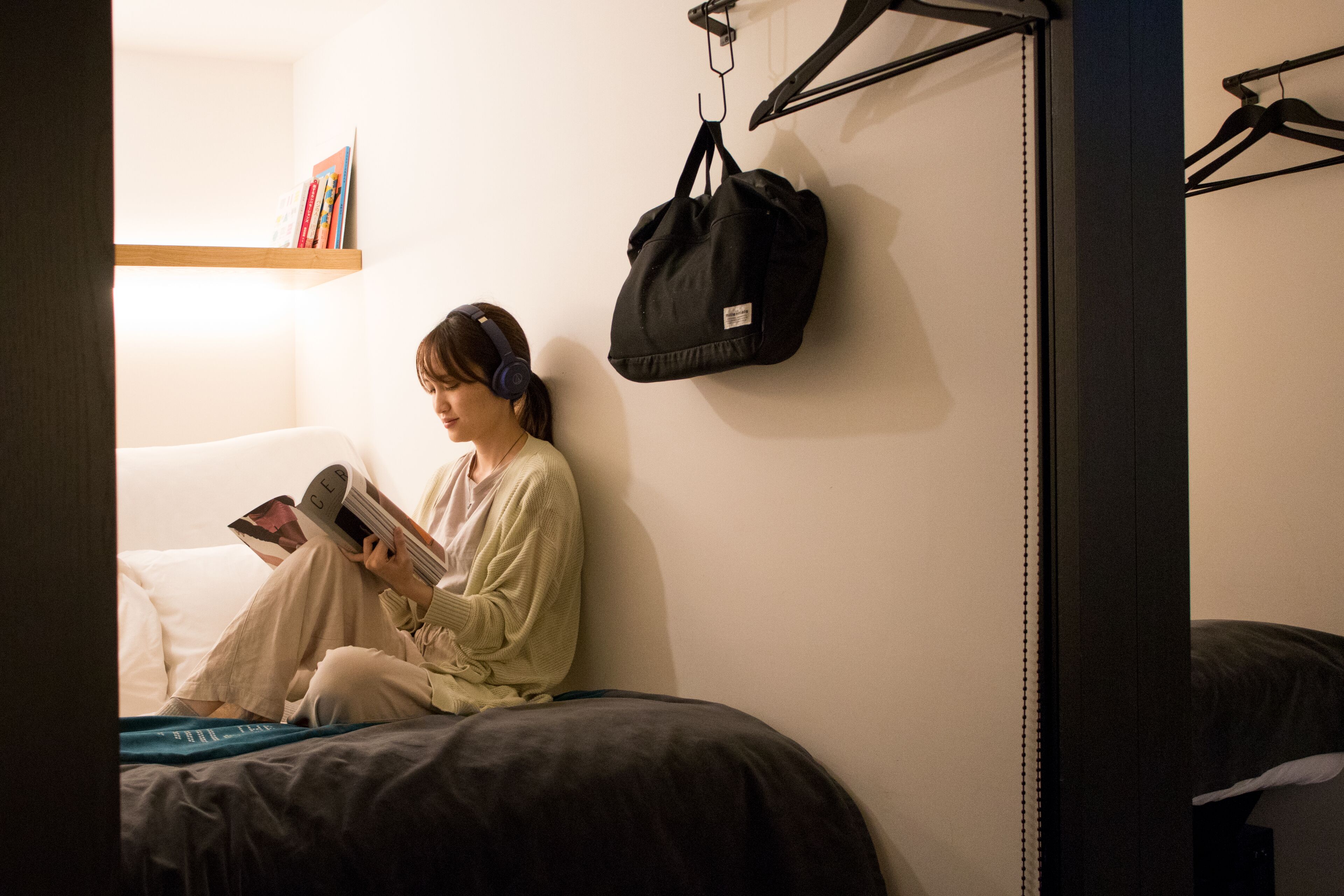 A person sitting on a bed reading a book in a room with a hanging bag and a shelf with books.
