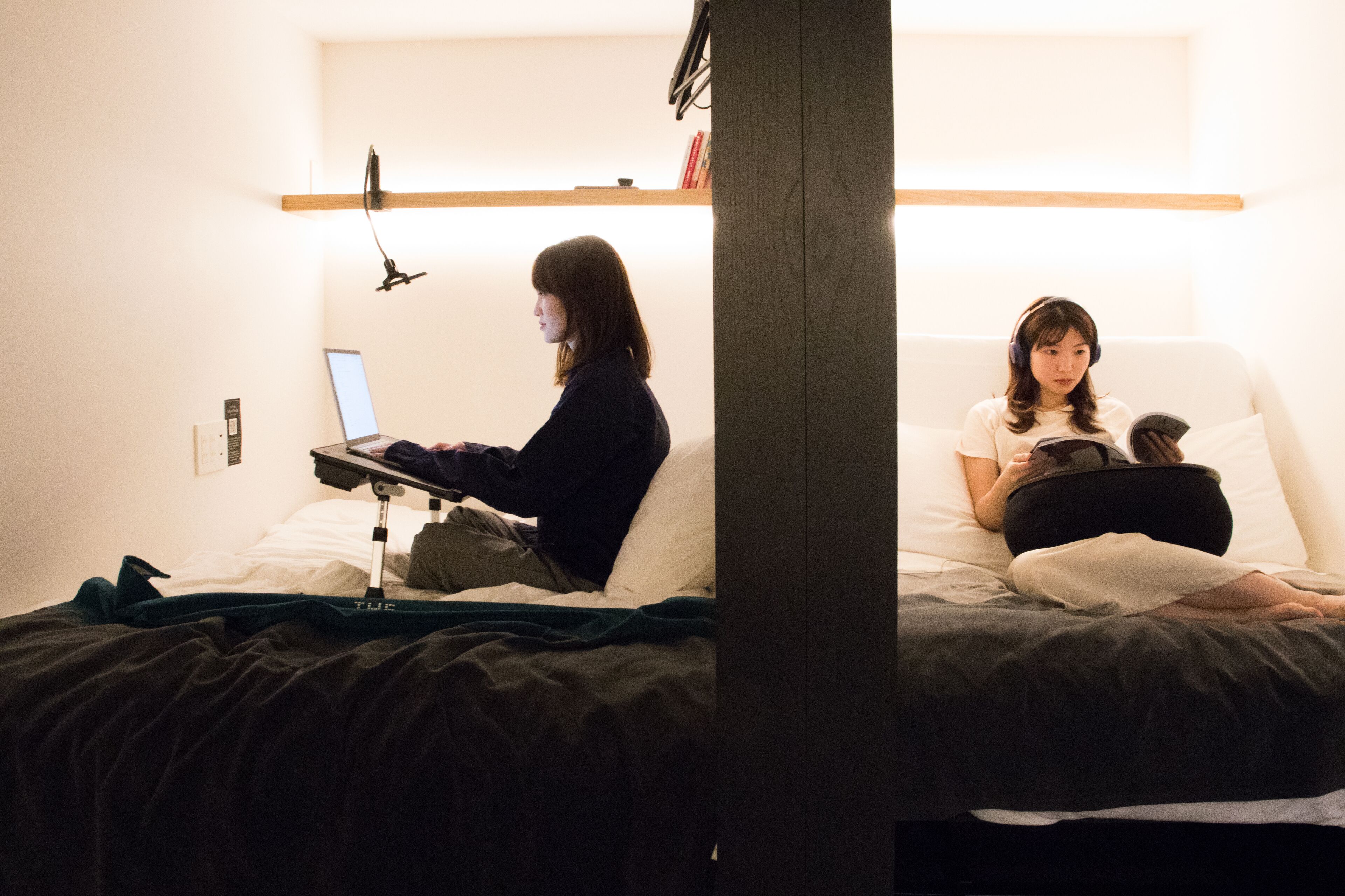 A room with a bed, a desk with a laptop, and a person reading a book.