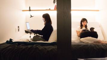 Una habitación con una cama, un escritorio con una computadora portátil y una persona leyendo un libro.
