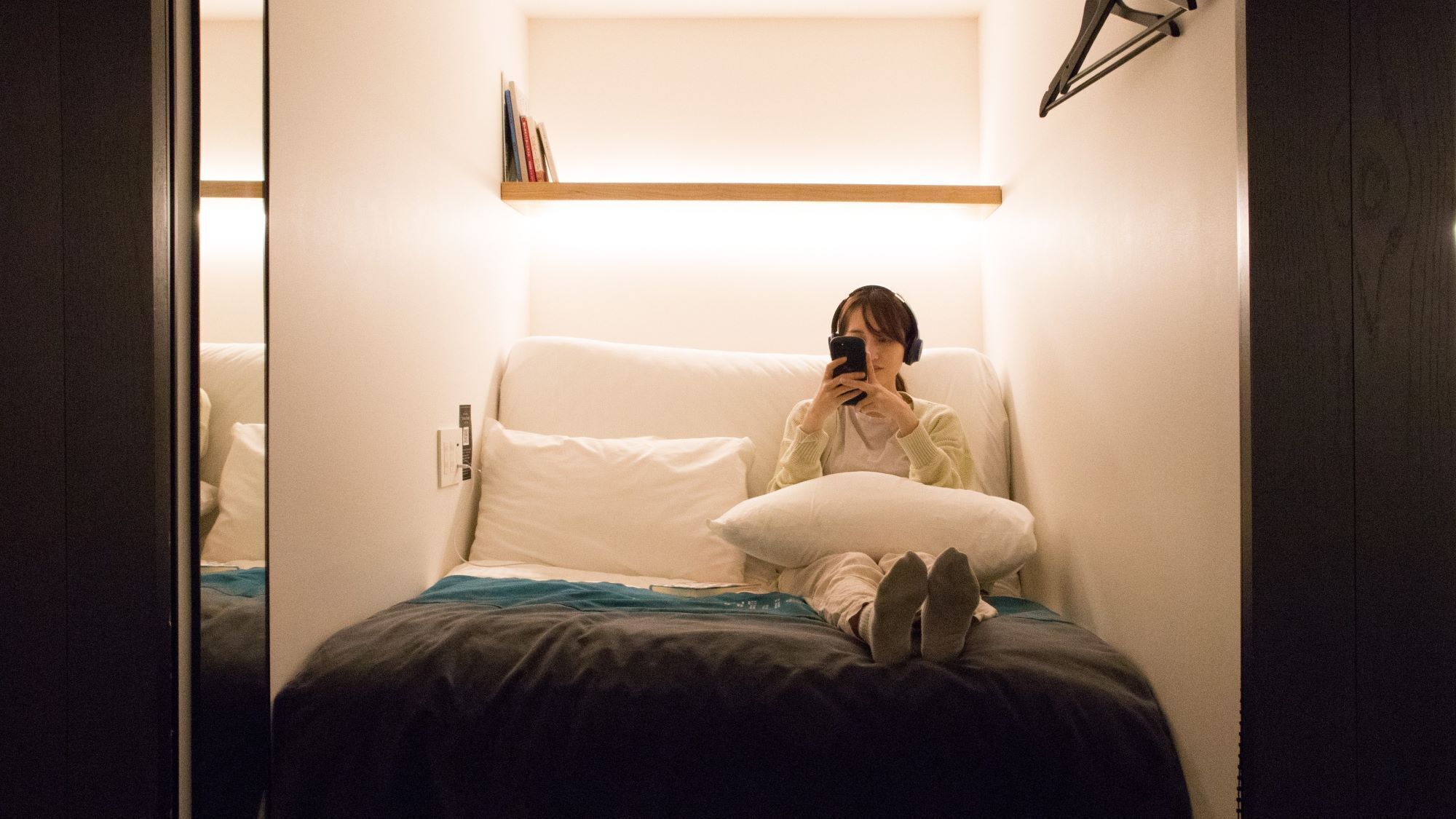 Una persona sentada en una cama en una habitación con pared blanca y estante de madera arriba.