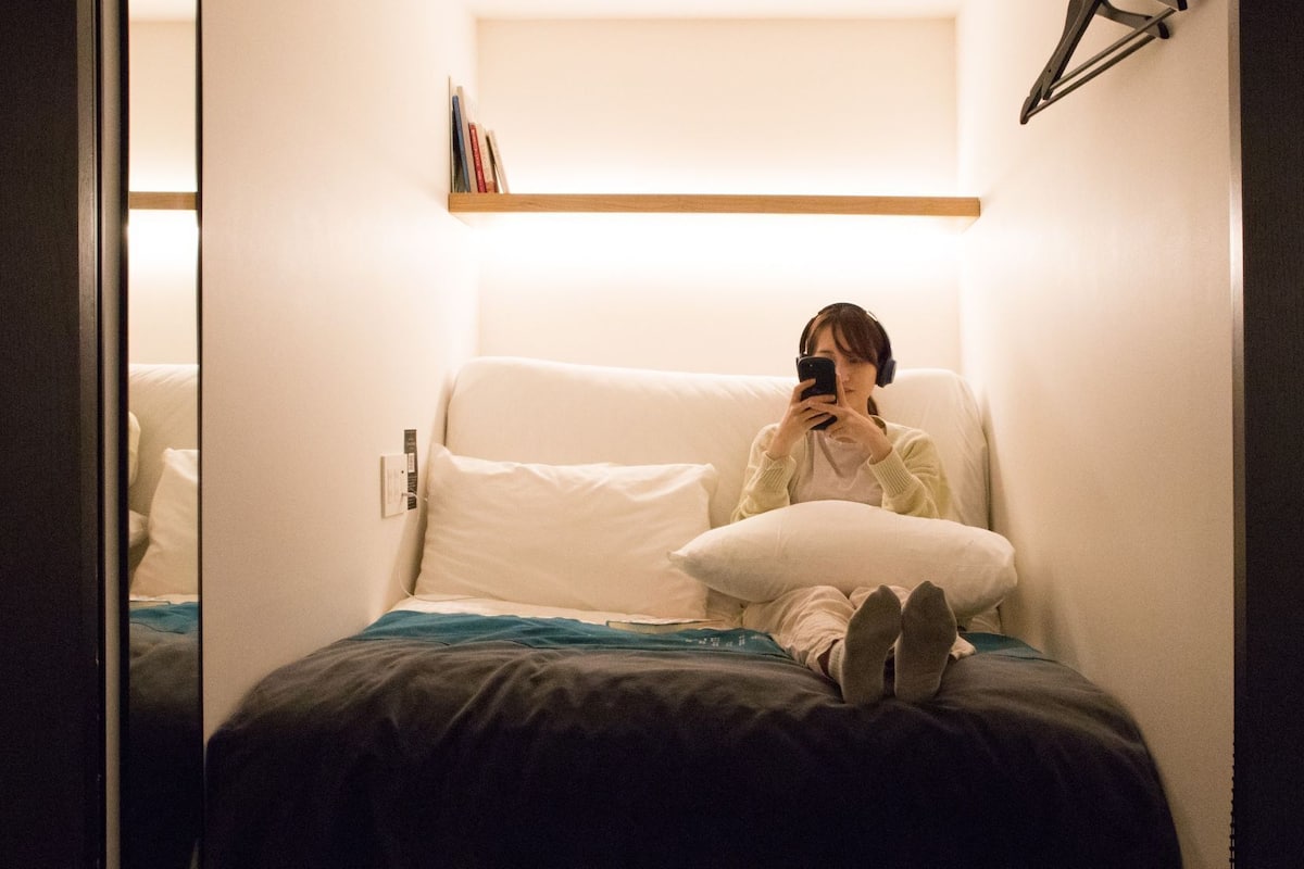 Uma pessoa sentada em uma cama em um quarto com parede branca e prateleira de madeira acima.