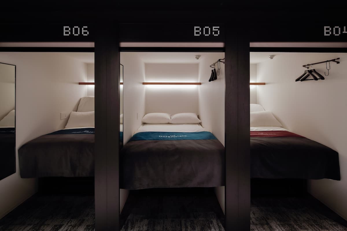 Quarto de hotel com três camas identificadas como B06, B05 e B04, cada uma com mesa de cabeceira e abajur.