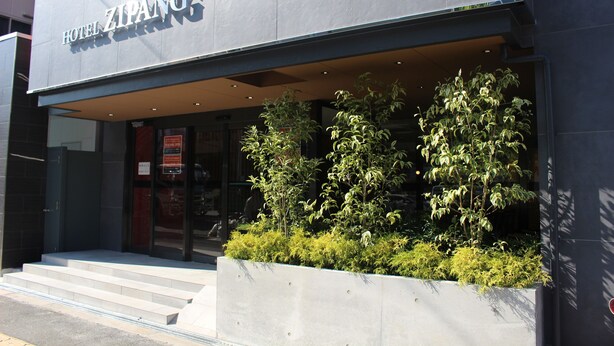 A hotel entrance with a modern facade, a visible staircase leading to the entrance, and a planter with greenery in front. - Hotel Zipang (Osaka)