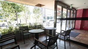 A modern indoor seating area with a view of the street, featuring round tables and chairs, and a mix of indoor and outdoor plants. - Hotel Zipang (Osaka)