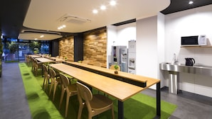 A modern dining area with a long wooden table and chairs, a kitchen area with a microwave and sink, and a green carpeted floor.
