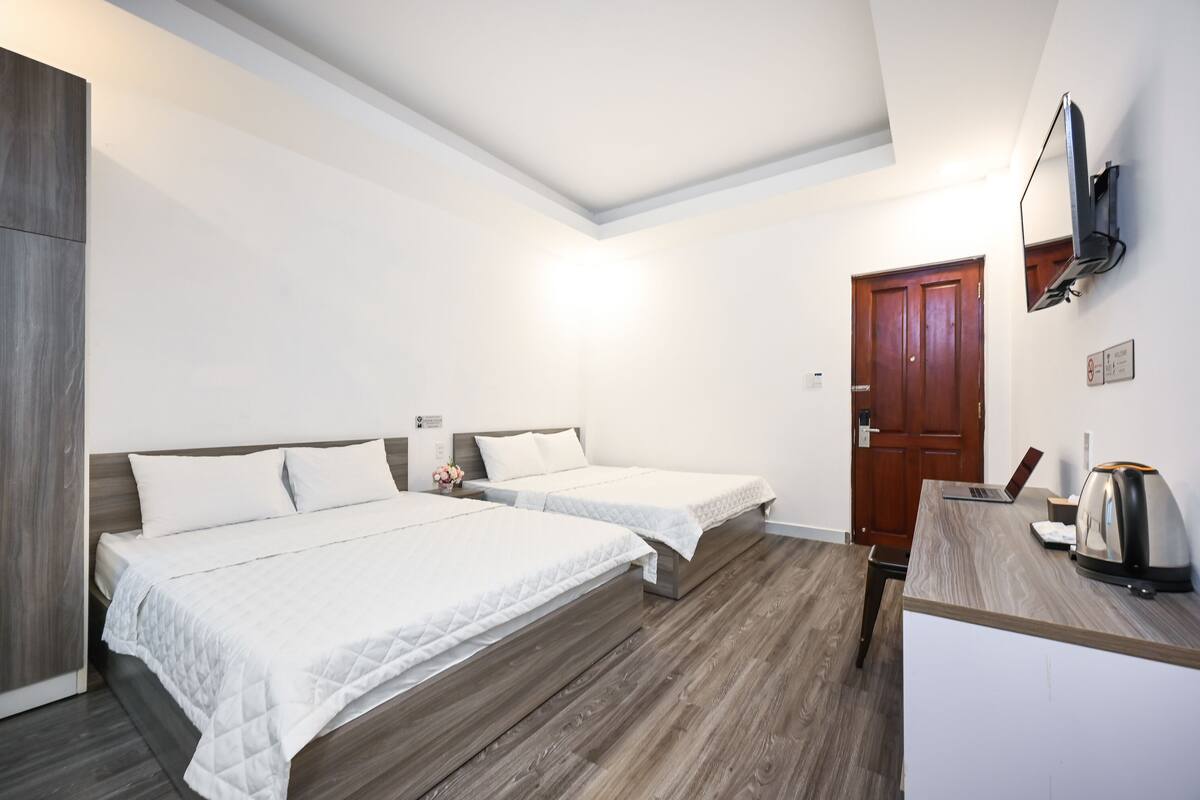 Habitación de hotel con dos camas, un escritorio con una computadora portátil y un hervidor, un armario de madera y un televisor montado en la pared.