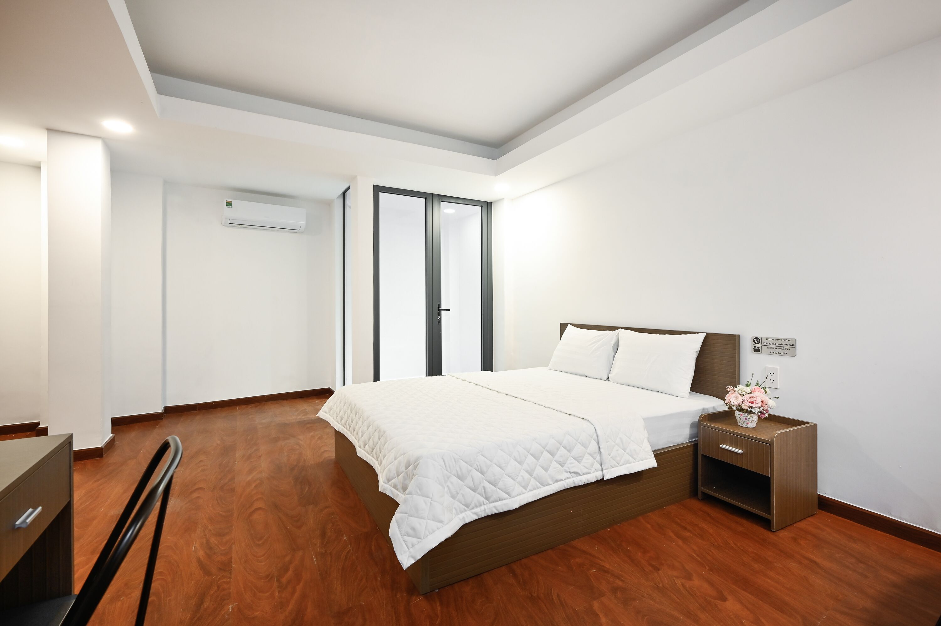 A hotel room with a bed, a nightstand, a desk, and an air conditioning unit.
