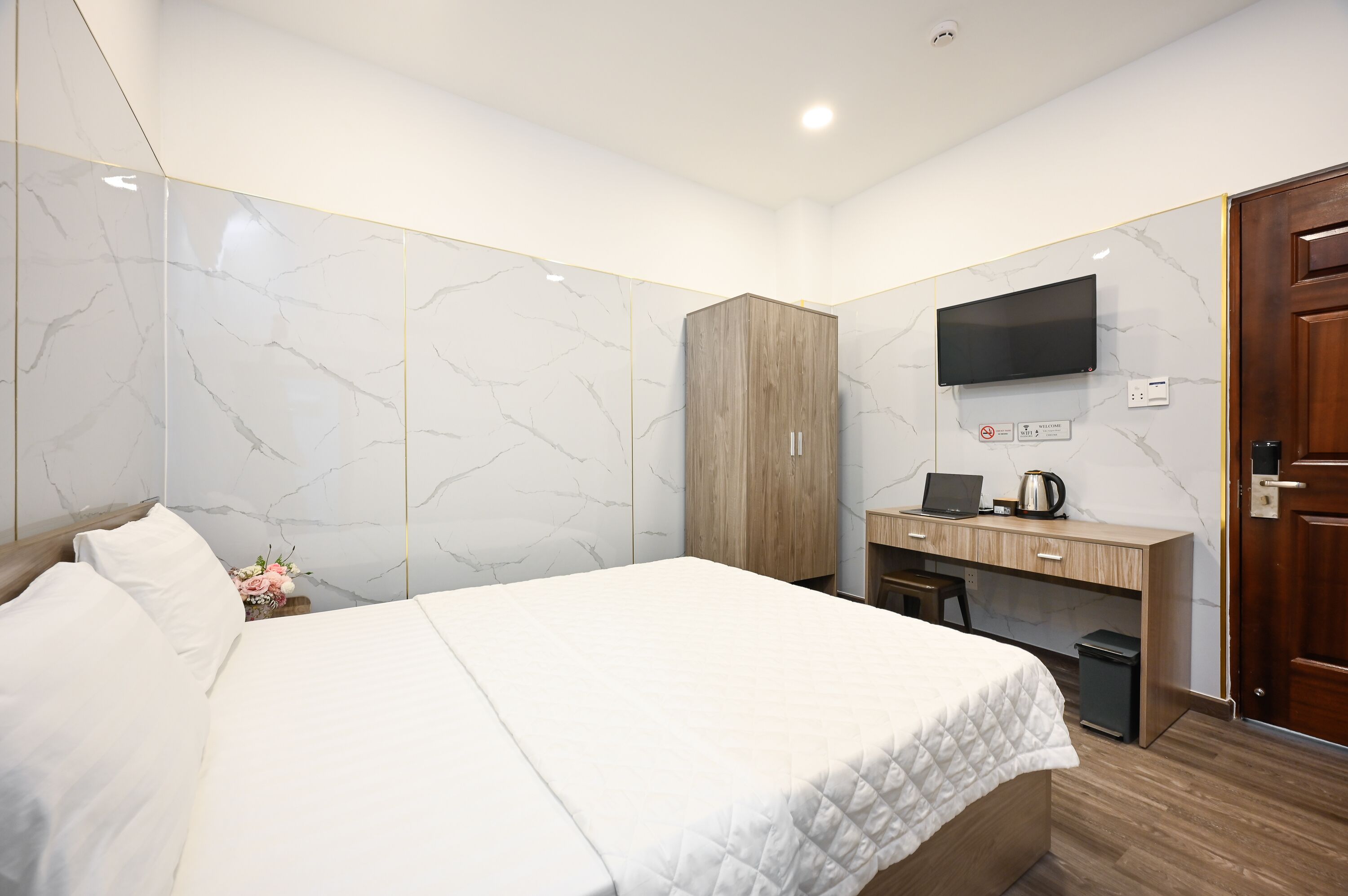 A modern hotel room with a large bed, a wooden wardrobe, a desk with a laptop, and a television mounted on the wall.