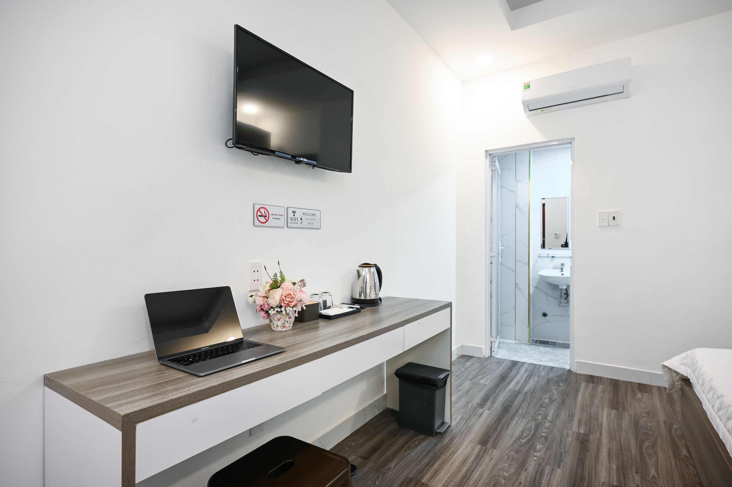 Una habitación de hotel moderna con un televisor de pantalla plana montado en la pared, una computadora portátil sobre un escritorio de madera, un cesto de basura y un bote pequeño para desechos.