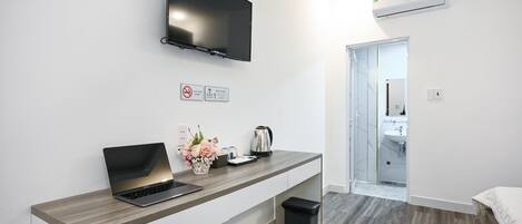 Una habitación de hotel moderna con un televisor de pantalla plana montado en la pared, una computadora portátil sobre un escritorio de madera, un cesto de basura y un bote pequeño para desechos.